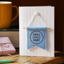 Personalised Father’s Day card with blue felt banner reading “Theo’s Lovely Daddy”. The handmade keepsake card with wooden dowel and button detail stands on a wooden table beside a yellow mug, book, gift, and pencil.