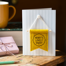 Greeting card with yellow banner and 'Henry's Lovely Daddy' text on a wooden surface with a mug and gift box.