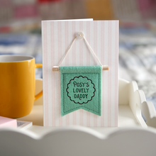 Personalised Father’s Day card with green felt banner reading “Posy’s Lovely Daddy”. The handmade keepsake card with wooden dowel and button detail stands on a wooden breakfast tray beside a yellow mug, book, and gift.
