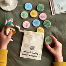 Hands holding a small toy and a bag labeled 'Henry & Daddy's Memory Making Ideas' with wooden activity tokens on a green table.