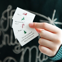 A child wearing a deep green jumper is holding tiny Christmas wish cards with festive icons, perfect as DIY Christmas cracker fillers or stocking stuffers.