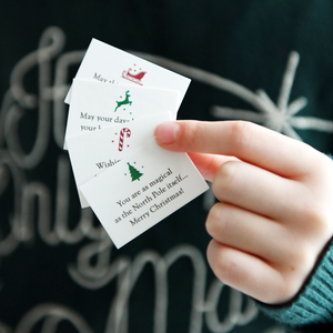 A child wearing a deep green jumper is holding tiny Christmas wish cards with festive icons, perfect as DIY Christmas cracker fillers or stocking stuffers.