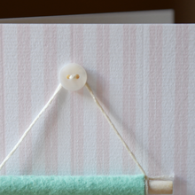 A striped card with a mother of pearl button stitched onto it, with a tiny felt banner hanging below.
