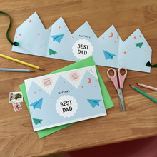 A flat lay image of a personalised Best Dad Father's Day greetings card with a blue crown design lays on a green envelope. Beside it is the Father's Day crown that has been made from the card. Both rest on a wooden desk with pastel coloured pencils, scissors, and vintage stamps around the cards.