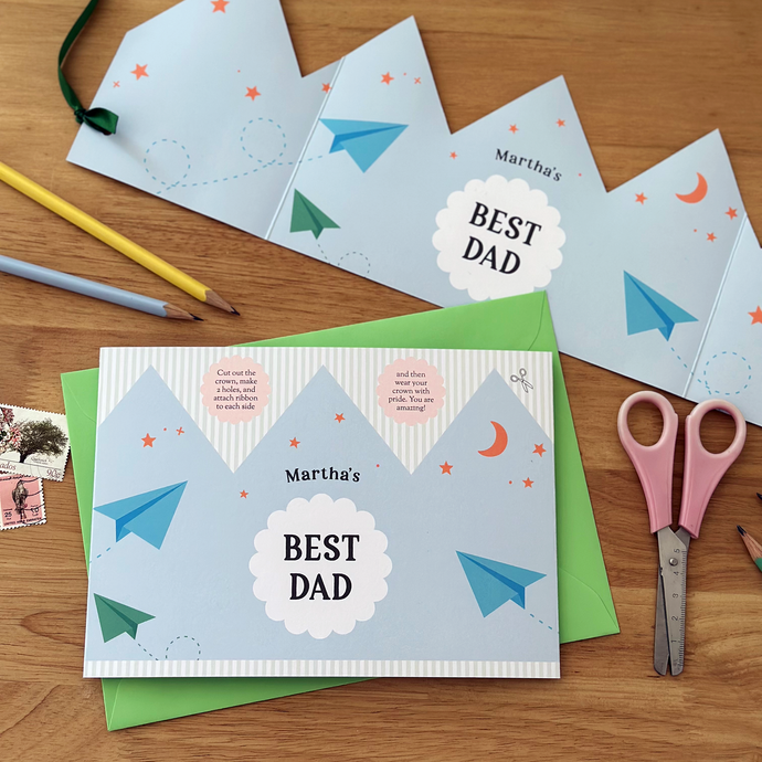 A flat lay image of a personalised Best Dad Father's Day greetings card with a blue crown design lays on a green envelope. Beside it is the Father's Day crown that has been made from the card. Both rest on a wooden desk with pastel coloured pencils, scissors, and vintage stamps around the cards.