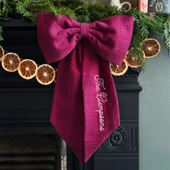 A handmade Christmas bow made from deep burgundy cotton with the words "The Clempsons" on one tail is hung from a black mantlepiece that is decorated for Christmas with greenery, orange slice garland, and candles.
