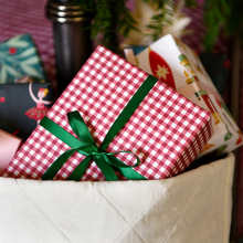 A wrapped gift box with red and white gingham paper and a green ribbon bow, nestled in a white quilted basket with other wrapped presents in the background.