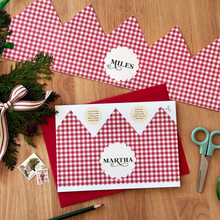 A personalised Christmas crown card with a red gingham design lays on a red envelope, on a wooden table.