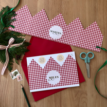 A personalised Christmas crown card with a red gingham design lays on a red envelope, on a wooden table. 