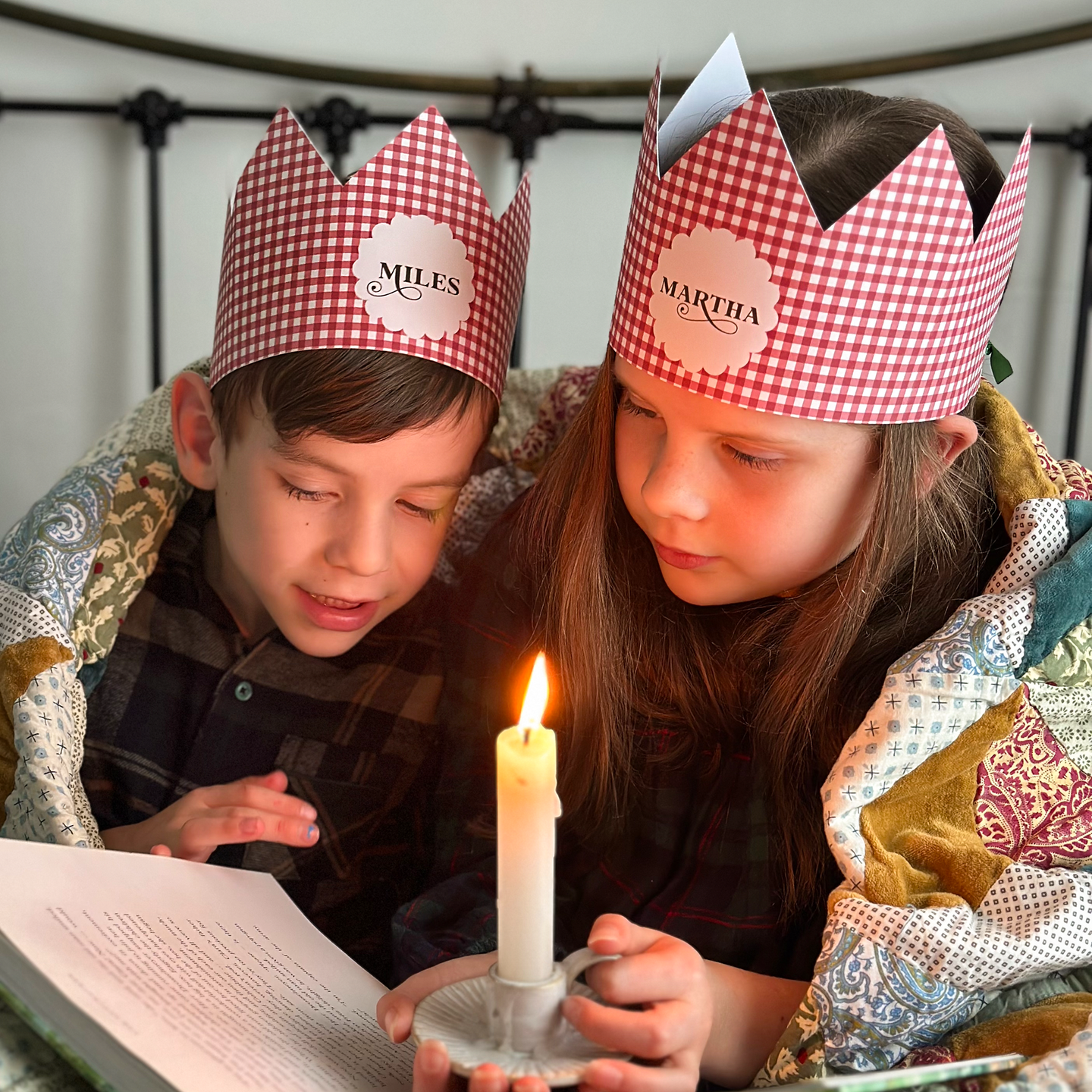 Two children wearing red gingham personalised Christmas crowns are wrapped in a cosy patchwork quilt reading a book together by candlelight.