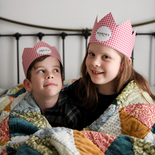 Two children wearing red gingham personalised Christmas crowns are wrapped in a cosy patchwork quilt giggling together.