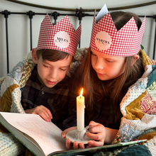 Two children wearing red gingham personalised Christmas crowns are wrapped in a cosy patchwork quilt reading a book together by candlelight.