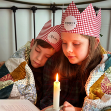 Two children wearing red gingham personalised Christmas crowns are wrapped in a cosy patchwork quilt reading a book together by candlelight.