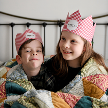 Two children wearing red gingham personalised Christmas crowns are wrapped in a cosy patchwork quilt giggling together.
