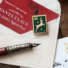 A postage stamp-style enamel pin featuring a gold reindeer on a dark green background with the number 25, displayed on a cream fabric surface with a red Santa-themed card, a decorative pen, and part of a letter visible.
