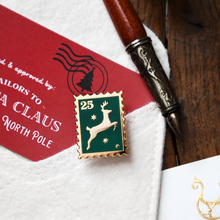 A Christmas-themed postage stamp enamel pin with a green background, gold border, and a white jumping reindeer, styled on a white envelope next to a red holiday card and a wooden pen on a dark wood surface.