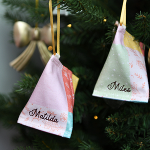 A handmade fabric fortune cookie Christmas decoration in pastel patchwork print, personalised with the name “Matilda” in brown, hangs from a golden ribbon on a Christmas tree branch.