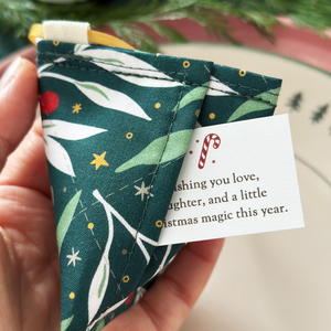 A handmade reusable fortune cookie Christmas decoration made from green festive foliage fabric is being held with a tiny Christmas wish card peeking out from the hidden pocket inside.