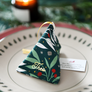 A green festive foliage fabric fortune cookie with gold personalised name “Theo” sits on a Christmas dinner plate, ready as a handmade festive place setting with a hidden Christmas wish card beneath.