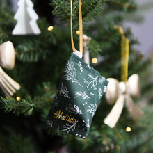 A handmade fabric fortune cookie Christmas decoration in green festive foliage print, personalised with the name “Matilda” in gold, hangs from a golden ribbon on a Christmas tree branch.