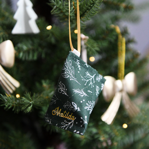 A handmade fabric fortune cookie Christmas decoration in green festive foliage print, personalised with the name “Matilda” in gold, hangs from a golden ribbon on a Christmas tree branch.