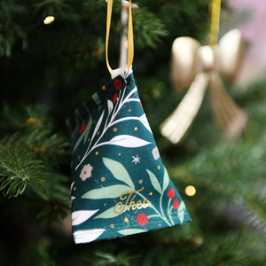 A handmade fabric fortune cookie Christmas decoration in green festive foliage print, personalised with the name “Theo” in gold, hangs from a golden ribbon on a Christmas tree branch.
