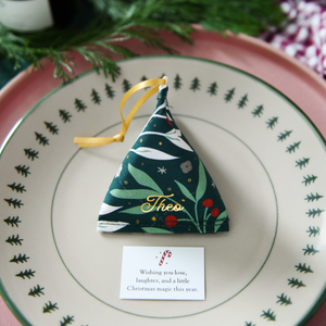 A green festive foliage fabric fortune cookie with gold personalised name “Theo” sits on a Christmas dinner plate, ready as a handmade festive place setting with a hidden Christmas wish card beneath.