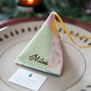 A pastel patchwork fabric fortune cookie with gold personalised name “Miles” sits on a Christmas dinner plate, ready as a handmade festive place setting with a hidden Christmas wish card beneath.