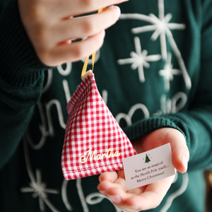 A child wearing a green Christmas jumper holds a red gingham personalised fabric fortune cookie with the name “Martha” in gold, revealing a tiny wish card inside that reads “You are as magical as the North Pole itself… Merry Christmas!”