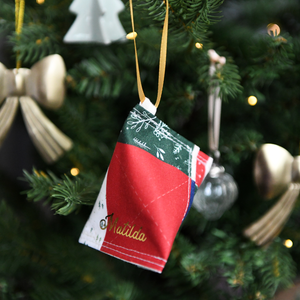 A handmade fabric fortune cookie Christmas decoration in green festive patchwork print, personalised with the name “Matilda” in gold, hangs from a golden ribbon on a Christmas tree branch.