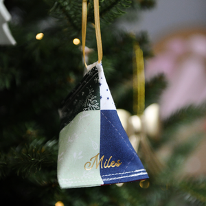 A handmade fabric fortune cookie Christmas decoration in a traditional patchwork print, personalised with the name “Miles” in gold, hangs from a golden ribbon on a Christmas tree branch.