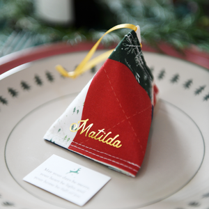 A traditional patchwork design fabric fortune cookie with gold personalised name “Matilda” sits on a Christmas dinner plate, ready as a handmade festive place setting with a hidden Christmas wish card beneath.