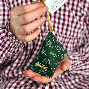 A child wearing a red gingham dress holds a green greenery personalised fabric fortune cookie with the name “Matilda” in gold, revealing a tiny wish card inside that reads “You are the magical sparkle in this season's story… Merry Christmas!”