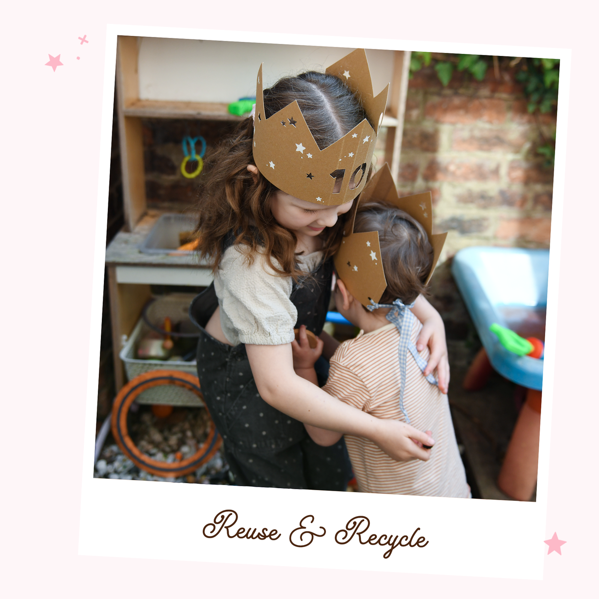 Two young children wearing neutral clothes and handmade cardboard crowns hug each other in front of a mud kitchen.