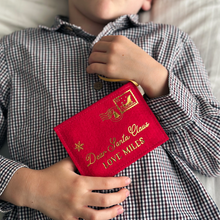 A child wearing a gingham shirt is holding a handmade red felt Christmas envelope that reads "Dear Santa Claus, Love Miles" in gold writing with a golden sleigh stamp in the top right hand corner.
