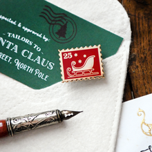 A square red Christmas-themed enamel pin shaped like a postage stamp, featuring a white sleigh and stars with a gold border, displayed on a white felt pocket knife next to a green card and a silver fountain pen nib.