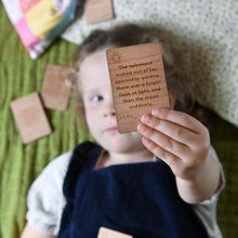 A young child is lying on a bed, covered with a patchwork quilt, reading wooden story prompt cards from a personalised 'Bedtime Stories' bag, designed for storytelling with Daddy.