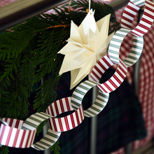 A close-up of a hanging paper chain garland with red, white, and green striped loops, alongside a cream-colored folded paper star ornament, displayed against dark green pine branches.