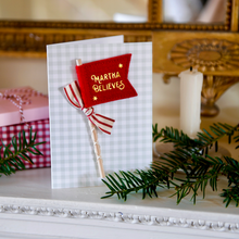 A handmade Christmas greeting card with a red felt flag reading 'MARTHA BELIEVES' and a striped ribbon bow, displayed on a white mantelpiece with holiday greenery and a candle.