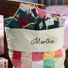 A tiny Christmas wish card is tucked into a wrapped Christmas gift that is one of a pile of gifts peeking out of a patchwork Christmas sack.