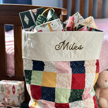 A tiny Christmas wish card is tucked into a wrapped Christmas gift that is one of a pile of gifts peeking out of a patchwork Christmas sack.
