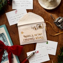 A cream-colored fabric Christmas stocking ornament or pouch with gold embroidery reading 'MABEL'S 24 TINY CHRISTMAS WISHES' and a postage stamp motif, styled on a wooden tabletop with handwritten cards, a red ribbon, and holiday greenery.
