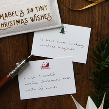 A styled flat-lay of handmade Christmas note cards on a dark wooden table, featuring a white fabric patch with gold embroidered text, a gold ribbon, a silver fountain pen, and green pine branches.