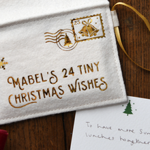 A close-up of a white felt Christmas ornament or pouch with gold embroidered text reading 'MABEL'S 24 TINY CHRISTMAS WISHES' and a gold postage-stamp style bell motif, placed on a wooden surface next to a handwritten note.