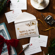 A white felt Christmas letter envelope with gold foil stamp details and the text 'MABEL'S 24 TINY CHRISTMAS WISHES' lies on a wooden tabletop surrounded by holiday-themed props, including handwritten cards, evergreen branches, a red ribbon, and a small mechanical device.