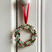 An heirloom Christmas wreath decoration with holly leaves and red berries hangs from a red ribbon on a white door handle.