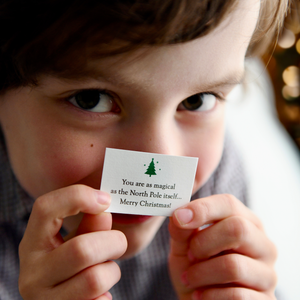 A young child holds a tiny Christmas wish card in front of his face. The card has a Christmas tree illustration and the words "You are as magical as the North Pole... Merry Christmas!"